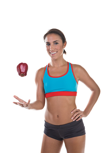 Portrait of a happy fit young woman in throwing apple. She is standing with her hand on hip. Beautiful and slim lady is in sports clothing. Cheerful fitness model is isolated on white background.