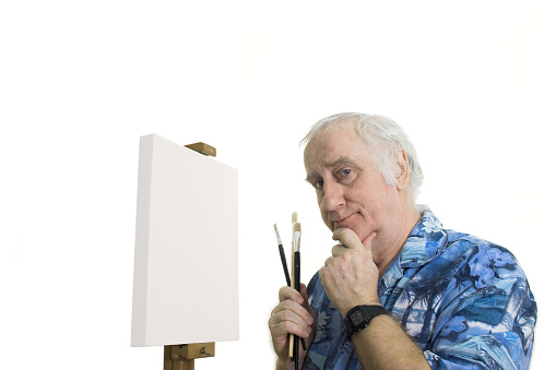senior artist waits quizzically in front of blank canvas on easel, for inspiration. 