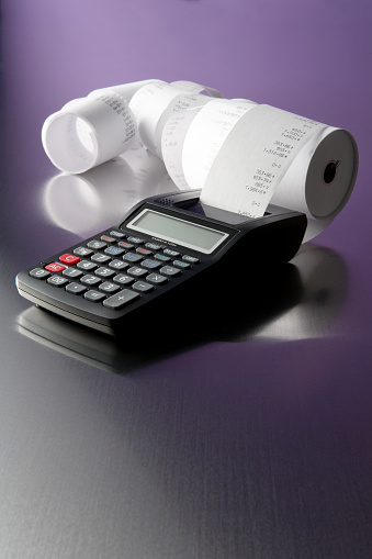 A black printing calculator with several rolls of printed receipts.  The calculator is shown against a reflective purple and gray background