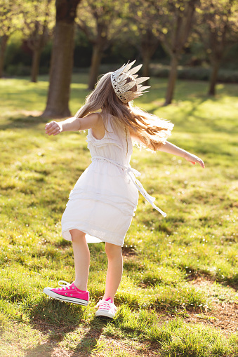 Little blond girl wearing a white dress, pink tennis shoes and a crown pretending to be a princess and dancing in the park.