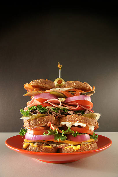 The Classic Dagwood Classic "Dagwood" (everything) sandwich, containing wheat bread, mustard, roast chicken, cheddar cheese, red onion, green leaf lettuce, tomatoes, bacon, mayonaise, pickles, roast beef, swiss cheese, salami, boloney, sardines, Canadian bacon, and a green olive. dagwood stock pictures, royalty-free photos & images