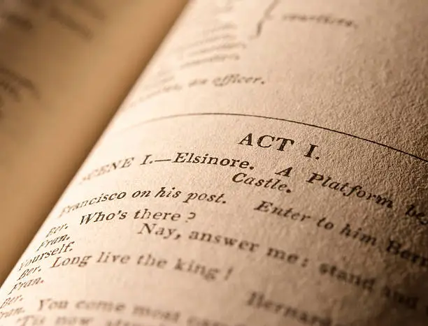 Side-lit, warmly toned macro shot of the first page of the play "Hamlet" in an antique edition (London 1839) of Shakespeare's Plays. Selective focus on the words "Act 1" and "Elsinore". The paper texture is clearly visible, particularly to the right of the image.