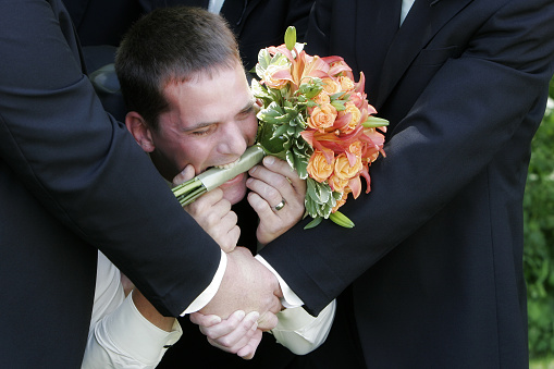 Happy groom lies across hands of his grooms men holding bride's bouquet in his mouth.