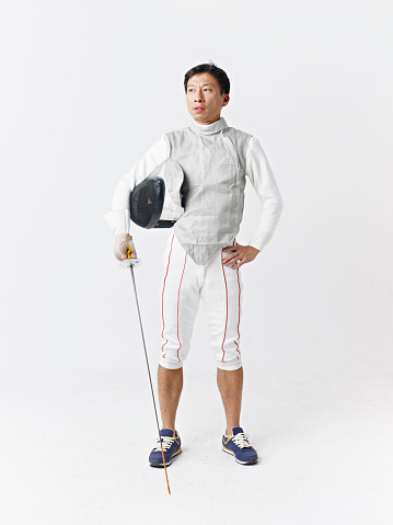 studio portrait of an male asian fencer with fencing mask and foil in hand, gray background.  