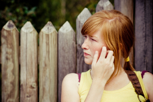 Beautiful red haired girl waiting near the wooden fence, looking aside and scratching her face.