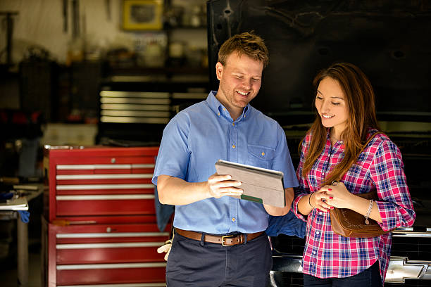 Happy customer discusses repairs with auto mechanic. Digital tablet. Happy customer.  A smiling Latin descent woman discusses automobile repairs with an automobile mechanic in a auto repair shop.  She is discussing the vehicle's problems with the mechanic, who is explaining results using his digital tablet.  They are in an automobile repair shop.  SUV-style vehicle. auto mechanic photos stock pictures, royalty-free photos & images