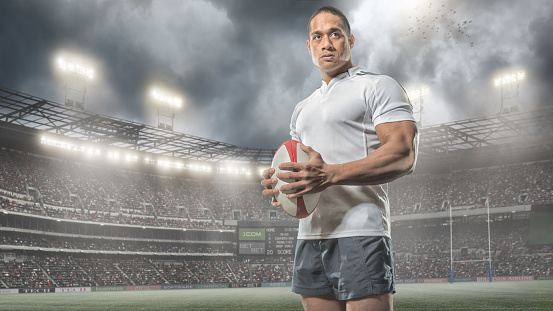 A low angle portrait of a professional male rugby player from waist upwards holding rugby ball and looking into the distance. The player is wearing a generic white rugby shirt and blue shorts and is standing in a generic outdoor floodlit rugby arena full of spectators under a stormy evening sky. 