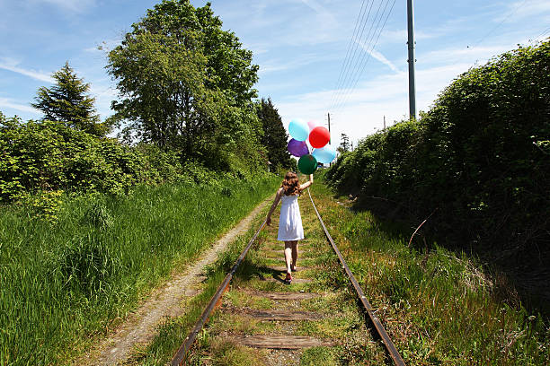 A little girl walking on the railroad tracks holding balloons.