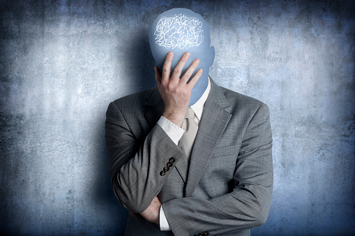 A man standing in a suit with his head in his hands.  The man's head is reduced to its simple shape, devoid of any detail, and is silhouetted against the background. A scribble design is located over his head representing the confusion and stress he is feeling.