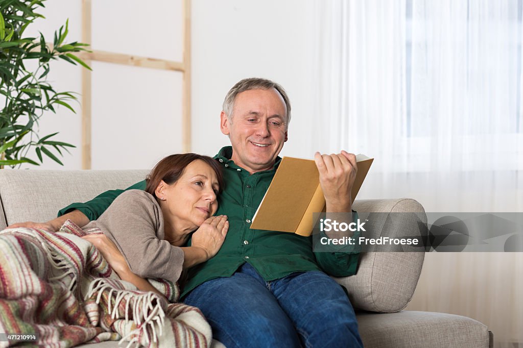 Mature couple relaxing on couch at home Happy mature adult couple with casual clothes sitting on a sofa in a modern style home, the mature man reading a book Couple - Relationship Stock Photo