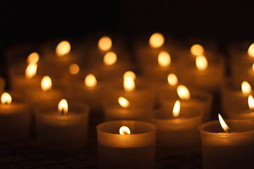 A large group of lit votive candles against a black background. Selective soft focus, with the sharpest point on the two glass jars in the foreground. Copy space in the black areas at the top and bottom of the frame.