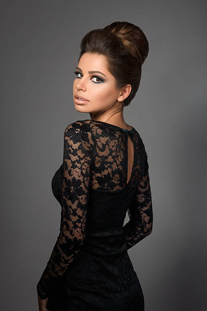 Beautiful young model in black dress with evening makeup and hairdo standing back to camera and looking back over her shoulder