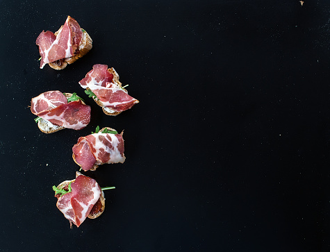 Bruschettas with dried tomatoes, arugula and smoked meat over a black backdrop with a copy space. Top view