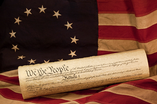 We the People, the opening words of the preamble to the Constitution of the USA, is prominent in this photograph of a rolled copy of the constitution. The parchment document is lying on an aged grungy colonial era American flag. This flag, popularly attributed to Betsy Ross, was designed during the American Revolutionary War and features 13 stars to represent the original 13 colonies.