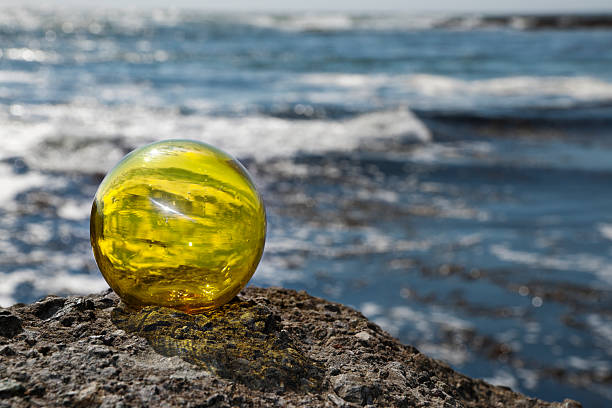 A yellow Japanese fishing float sits at the edge of a rocky ocean shoreline.
