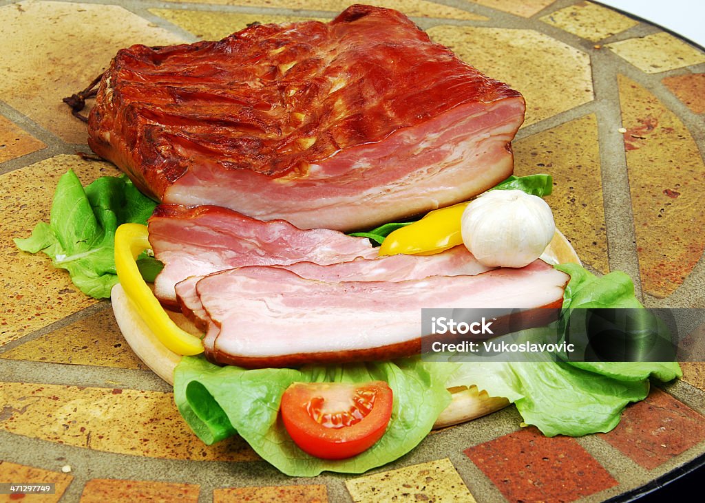 Sliced smoked bacon with wegetables. Big piece of a partialy sliced smoked bacon on the stone table. Appetizer Stock Photo