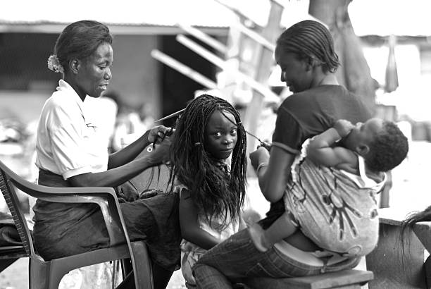 African Family Hairdressing Scene Accra, Ghana - December 21, 2009: African little girl has braids made by two women in the popular Osu district in Accra, Ghana. The younger woman is carrying a baby on her back. braided hair photos stock pictures, royalty-free photos & images