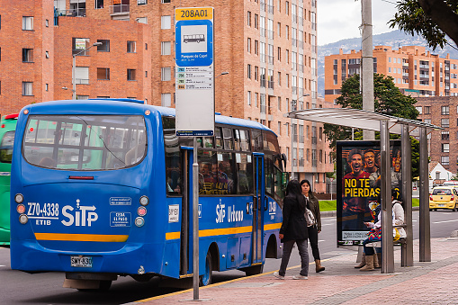 Bogota, Colombia - April 22, 2015: It is a no-car day in the Andean capital city of Bogota, Colombia in South America.  Only busses and taxis are allowed on the roads; Carrera 9, the avenue, is fairly free of traffic.  A few ladies are seen waiting for their bus. A bus has just pulled up at the stop. In the far background are the always present Andes Mountains. Photo shot in the afternoon sunlight; horizontal format.
