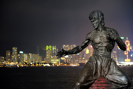 Hong Kong, China - November 14, 2009: The Bruce Lee statue in Hong Kong is a memorial figure of martial artist deceased in 1973. The bronze statue was erected on the Avenue of Stars. Hong Kong\\'s skyline with illuminated buildings and sea in background. Night time, high iso, grain.