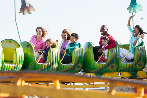 A multiracial group of people having fun on a rollercoaster ride at an amusement park.  The three adults and four children are smiling and the African American woman on the end is waving.