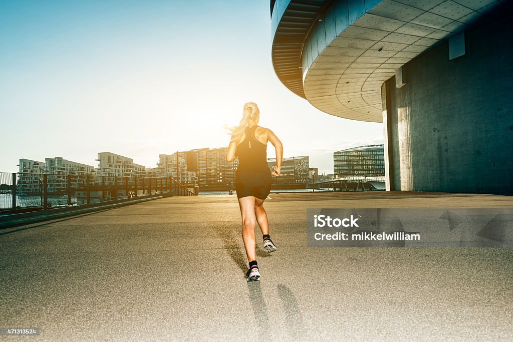 Sunset in the city and womans runs towards the horizon Female runner in the city while the sun sets. She is wearing sports clothes and has long blond hair. The photo shows her back as she runs towards the horizon where the sun is setting. City buildings to the side and in the background. 2015 Stock Photo