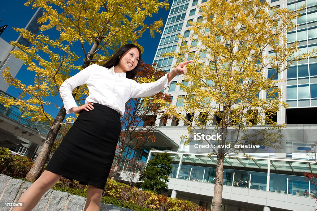 Confident Asian Female Executive Low angle 3/4 view of an attractive young adult woman in business attire standing with feet apart in a confident, "take charge" attitude.  With one hand on her hip  and the other pointing away, she shows a toothy smile as she looks in the same direction. She is dressed in business attire and is on a plaza outside a highrise in Tokyo. 25-29 Years Stock Photo