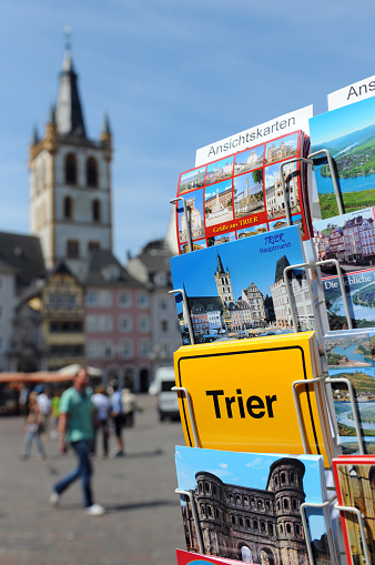 Trier, Germany - July 5, 2011: Tourists walking across the Market place in Trier centre (Germany) enjoying the sun. In Front typical postcard with town name Trier, Porta Nigra motive and Mosel river.