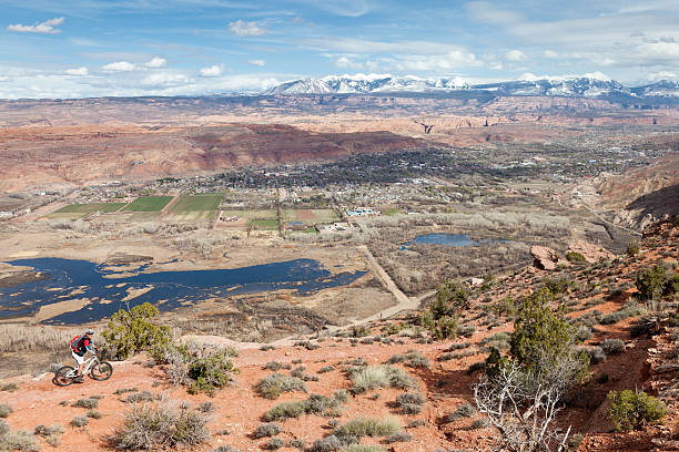 Moab viewpoint, Utah A male mountainbiker on his way on a single trail with beautiful view to the city of Moab, which is a city in Grand County, in eastern Utah, in the western United States. Moab hosts a large number of tourists every year, mostly visitors to the nearby Arches and Canyonlands National Parks. The town is a popular base for mountain bikers who ride the extensive network of trails including the famed Slickrock Trail, and off-roaders who come for the annual Moab Jeep Safari. In the background the snowcapped summits of La Sal Mountains are visible. la sal mountains stock pictures, royalty-free photos & images