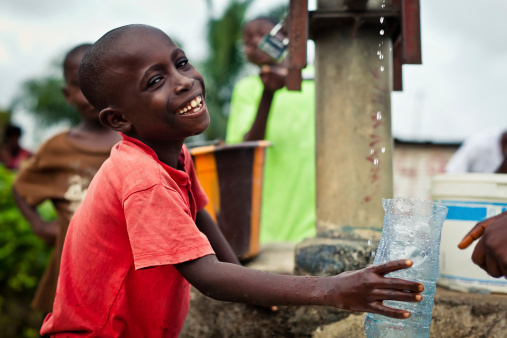 An African boy holding a funnel while clean water pours out of the spout of a water pump into a water jug.