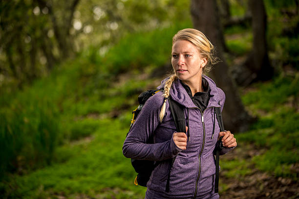 A young and attractive woman taking in the view of nature while carrying a backpack in the forest. She has a blond hair and a braid and is wearing a purple sweat shirt. Her hands are gripping her backpack straps. She is surrounded buy lush greens and brown colors of the forest.