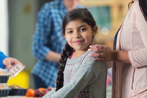 Elementary age Hispanic little girl is smiling while standing with her mom in line at food bank or soup kitchen. They are waiting to be served healthy meal by diverse group of volunteers. Mother's hands are resting on daughter's shoulders. 