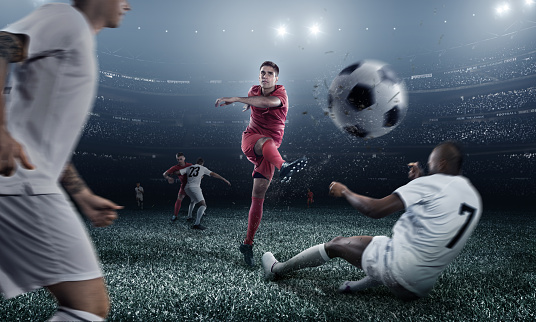 A male soccer player makes a dramatic play. He attempts to kick the ball with his feet. The opposite team players are going to block the ball, but fail. All players are wearing generic unbranded soccer uniform. The stadium is 3D rendered.