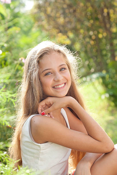 Portrait of  pretty teenage girl sitting, smiling, enjoying nature Portrait of  pretty teenage girl with long blond lush hair sitting outdoors in the park among the greenery at sunny day, posing, smiling, looking at camera. cute 15 year old girls stock pictures, royalty-free photos & images