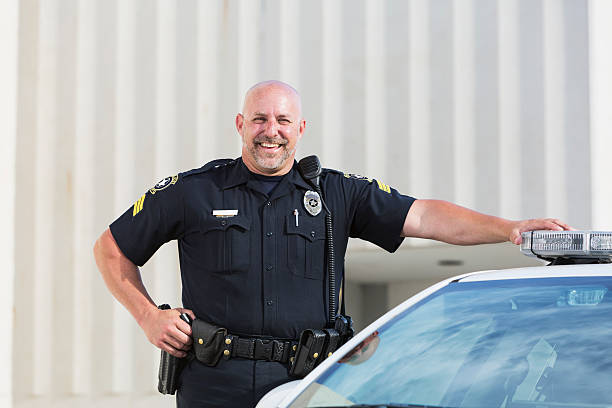 Happy police officer standing next to cruiser Policeman smiling, standing next to his police car looking at the camera. He is a heavyset man in his late 40s, with bald head. He has one hand on his hip and the other on top of the siren on his car. He is wearing a dark blue uniform. police force smiling officer stock pictures, royalty-free photos & images