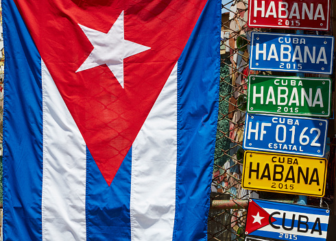 A Cuban flag hangs next to license plates on a chain link fence.