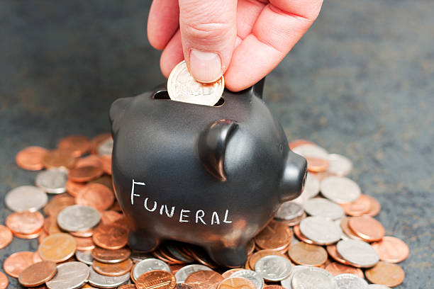 Funeral Piggy Bank on Coins A coin piggy-bank with "Funeral" written on it sitting on a pile of coins with a hand dropping a gold coin into the slot. funeral planning stock pictures, royalty-free photos & images