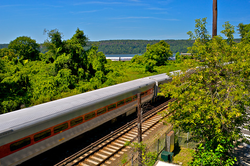 A Metro North commuter railroad train is seen in the foreground as an Amtrak long distance train in the background passes nearby the Hudson River at Spuyten Duyvil in the Bronx, New York City.  In the distance are the Palisades cliffs of New Jersey.