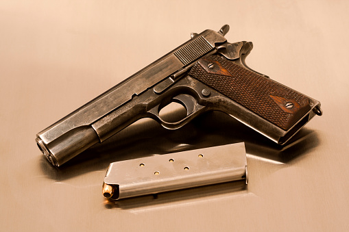 A US Government issue Colt 1911, semiautomatic handgun chambered for the .45 ACP cartridge with a loaded magazine on a brushed aluminum (stainless steel) background. This was the standard-issue side arm for the United States armed forces from 1911 to 1985. Markings on frame say 