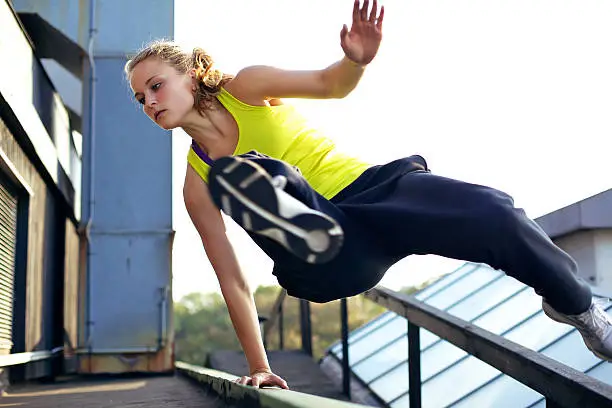 A woman traceur concentrating on vaulting over a railing on a high industrial building while demonstrating parkour.