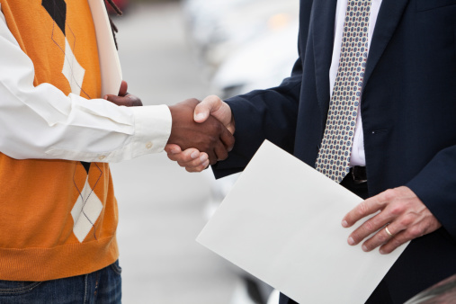 African American man in smart casual clothes shaking hands with Caucasian man in suit, standing in parking lot with row of cars in background.