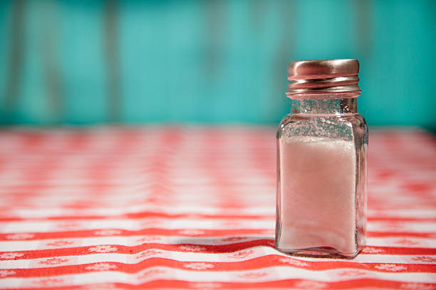One lone salt shaker filled with salt sits on top of a red checked tablecloth.  Outdoor summer picnic concept.  Blue background.  Restaurant or picnic table. Eat less sodium. Health concerns such as hypertension.  Copyspace to left.