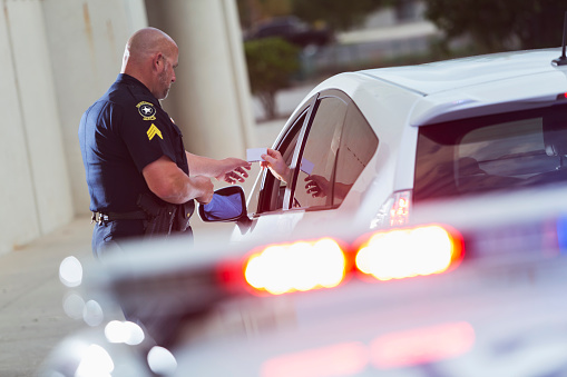 A police officer stands next to a car he pulled over for speeding,  reaching for the driver's identification card, which a man is handing him through the car window.  The police cruiser is out of focus in the foreground with lights flashing.