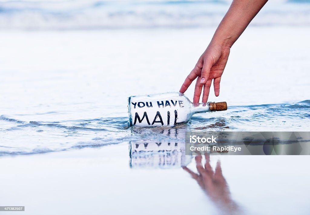 Modern message in bottle on beach says You have mail A hand reaches down for a bottle containing the message "you have mail" at the water's edge. The castaway seems to have internet access!  Message in a Bottle Stock Photo