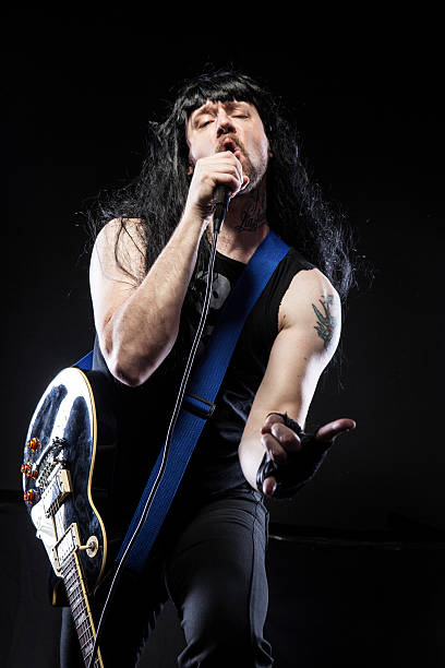 Rocker dude holding a mic with a guitar and black tank top.  Making a rocker hand signal in front of a black background.