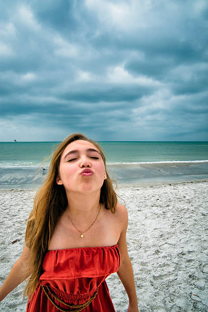 Beautiful girl wearing orange dress on the beach is kissing away the bad weather and the cloudy sky. This was taken in the Clearwater area, Florida.