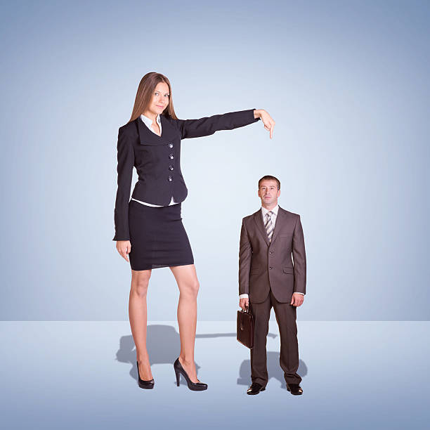 Smiling Young Businesswoman Pointing to small Businessman Smiling Young Businesswoman Pointing to small Businessman. Blue background tall person stock pictures, royalty-free photos & images