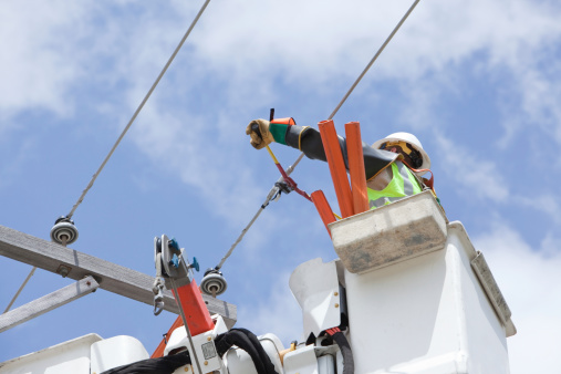 An electric utility lineman in a truck bucket is using a bolt cutter to cut a jumper wire connection on a high voltage power line. The worker is wearing insulated gloves and sleeves. The orange tubes are 