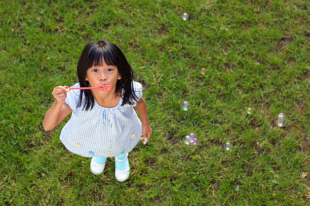 A cute little 6 year old Asian girl enjoys blowing bubbles into the air on a lovely summer day.