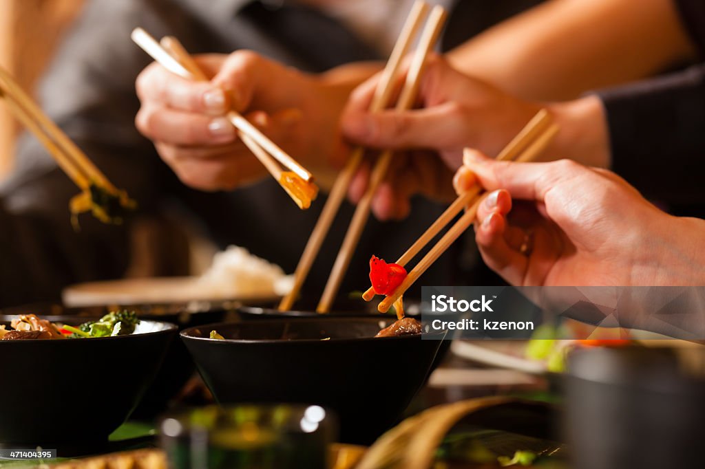 Young people eating in Thai restaurant Four sets of chopsticks grabbing Asian food out of various black bowls sitting on a table in a Thai food restaurant.  Three pairs of chopsticks have food between them, and one is in a black bowl.  There is a person's chest out of focus in the background.  There are four sets of chopsticks visible, but there are only three hands visible.  There is a set of chopsticks coming into view from the left.  The hand in front is in focus, while the two hands in the background are slightly blurred. Chinese Culture Stock Photo