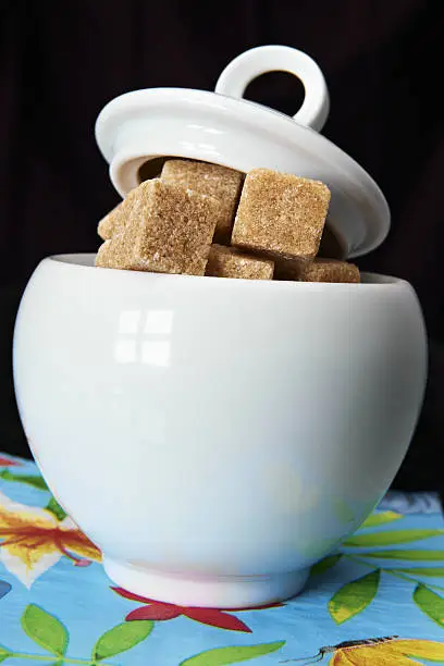 white sugarbowl with brown sugarcubes  and jars of coffee and sugar on mixed background: blue/flowery at the bottom and black above